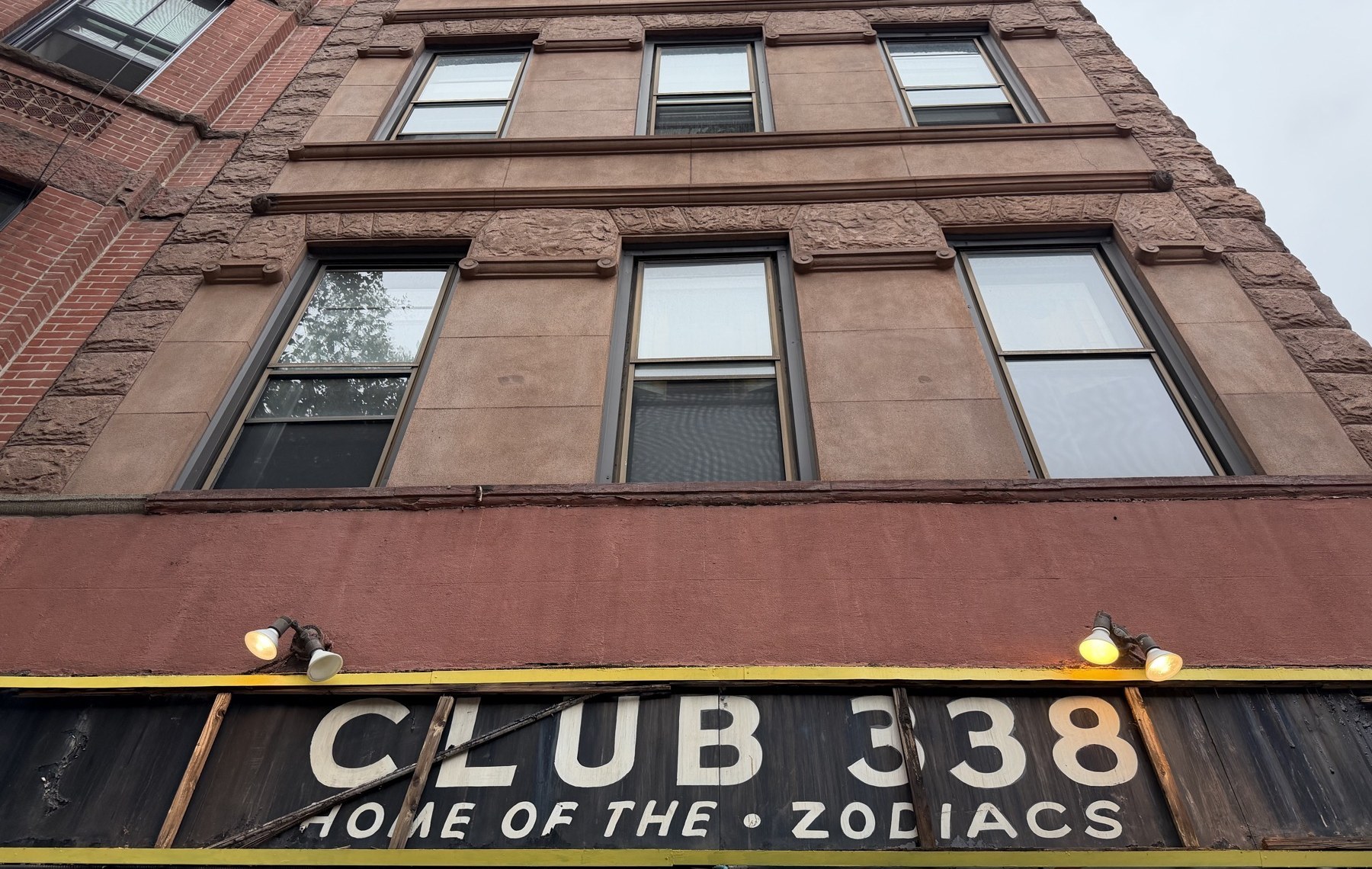 Low angle of the brick facade. A black board in a yellow frame runs across the lower third, with cream letters on it.